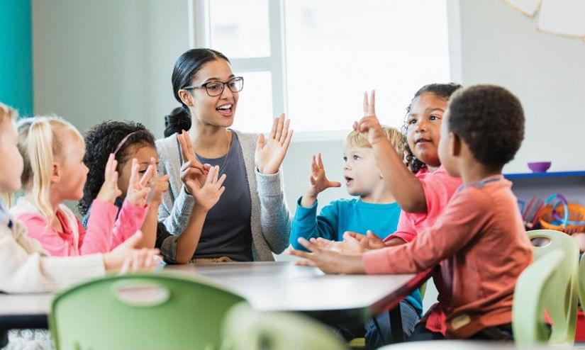 A teacher surrounded by young children with their hands up